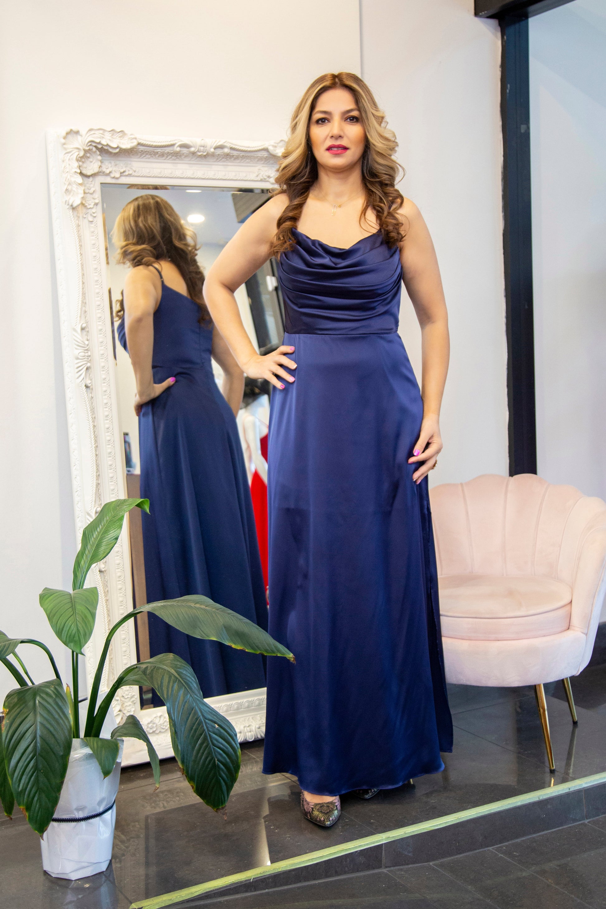 Woman in a blue dress standing in front of a mirror in a room with a plant and chair.