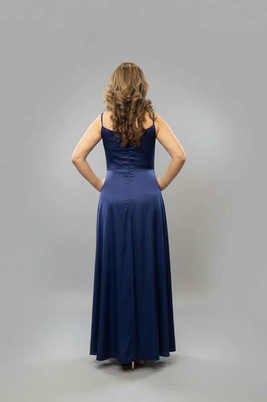 A woman stands with her back to the camera wearing a floor-length navy blue gown with thin straps and a fitted bodice, photographed against a plain light gray studio background.