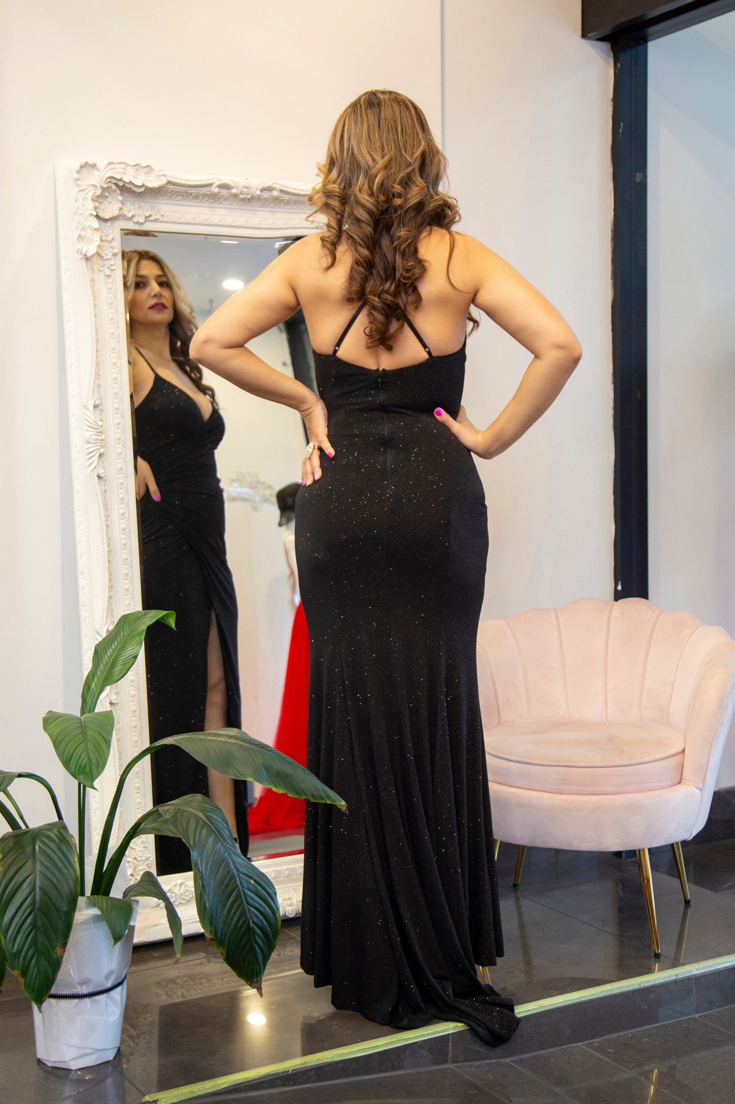 Woman in a black evening gown standing in front of a mirror in a room with a plant and chair.