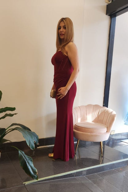 A woman wearing a sleeveless, floor-length burgundy evening gown stands in a boutique interior with a pink shell-shaped chair, a potted plant, and a window display.