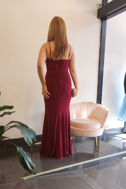 A woman stands with her back to the camera wearing a floor-length, sleeveless burgundy evening gown inside a boutique. A pink velvet chair, a potted plant, and a glass window displaying a mannequin are visible in the store.