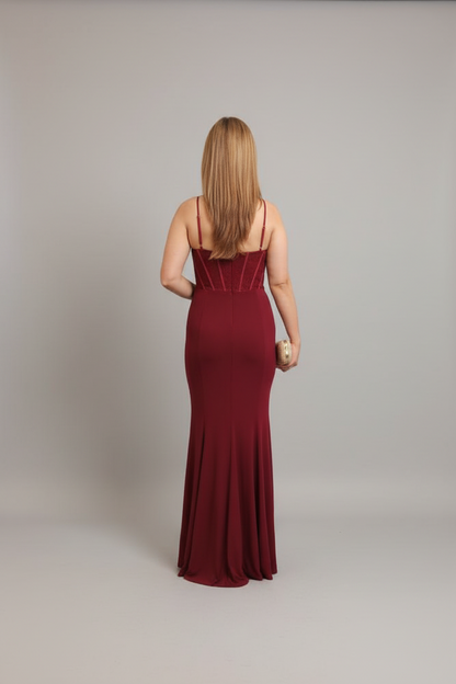 A woman viewed from behind wearing a floor-length burgundy maxi gown with thin straps and structured detailing at the back, standing against a plain light gray studio background.