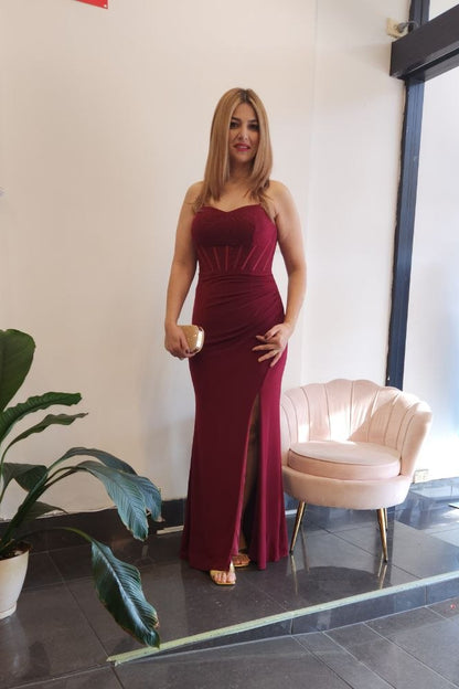 A woman stands indoors wearing a sleeveless, floor-length burgundy gown with a high front slit. The setting includes a dark tiled floor, a potted plant, a blush pink chair, and a glass door with a mannequin in a blue dress visible inside.