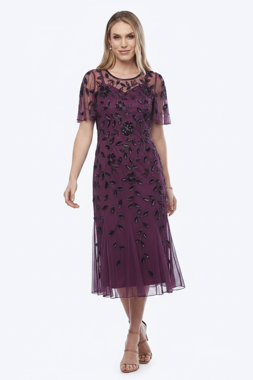 A woman modeling a short-sleeve midi dress in deep purple with black floral embroidery and sheer mesh sleeves, paired with nude high-heeled sandals against a plain white studio background.