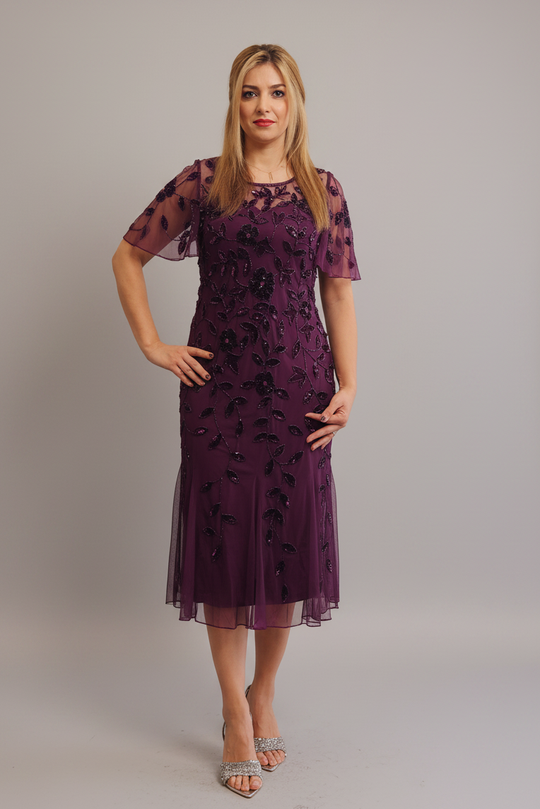 A woman modeling a knee-length, short-sleeved tulle dress in deep purple with black floral applique, standing against a neutral gray studio background.