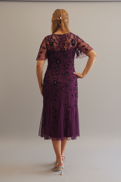 A woman wearing a short-sleeved burgundy tulle dress with black floral lace applique, standing in a room with a cream wall and a pink chair in the background.