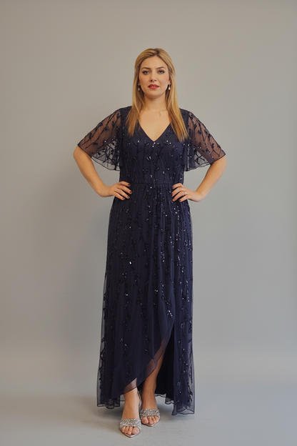 A woman modeling a navy blue lace and sequin maxi dress with sheer flutter sleeves and a high-low hem, standing against a neutral gray studio background.