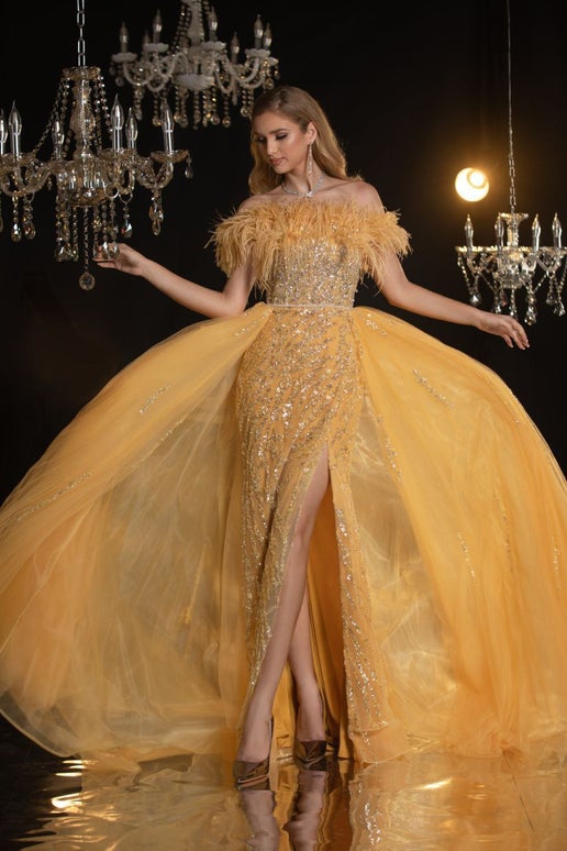Woman in a glamorous yellow evening gown with a high slit, standing under chandeliers.
