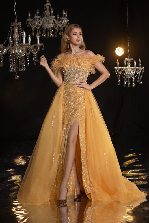 Woman in a glamorous yellow evening gown with a high slit, standing in a dimly lit room with chandeliers.