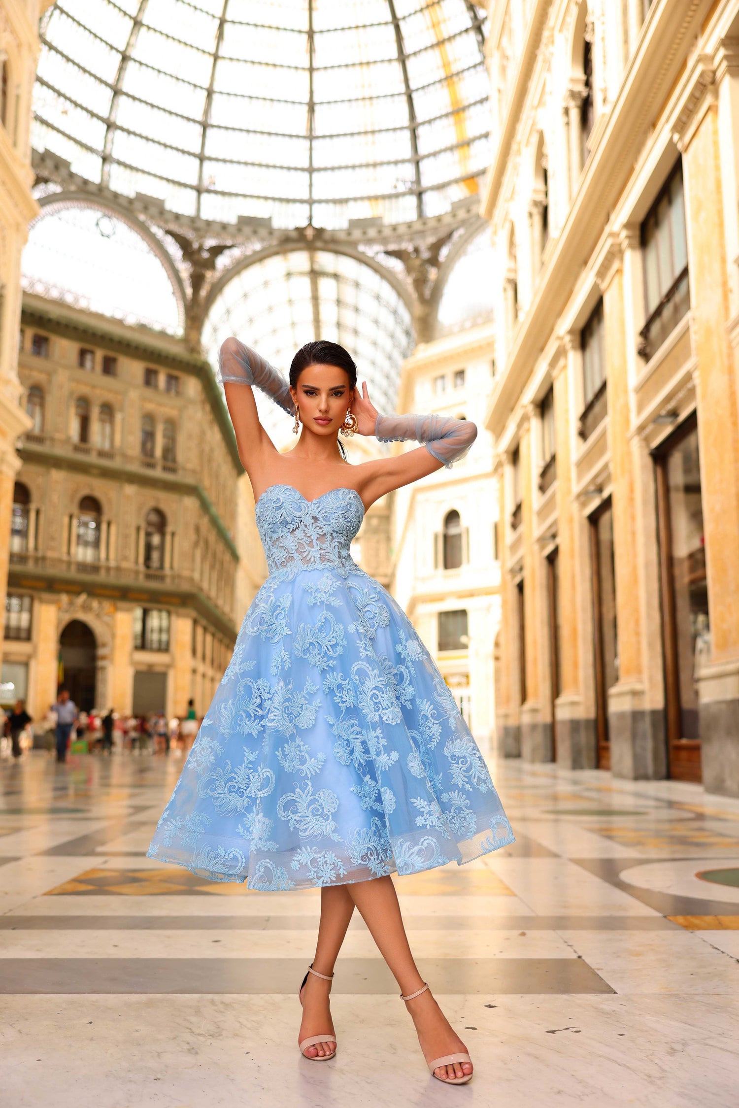 Woman in a light blue dress standing in an elegant indoor setting with architectural columns.