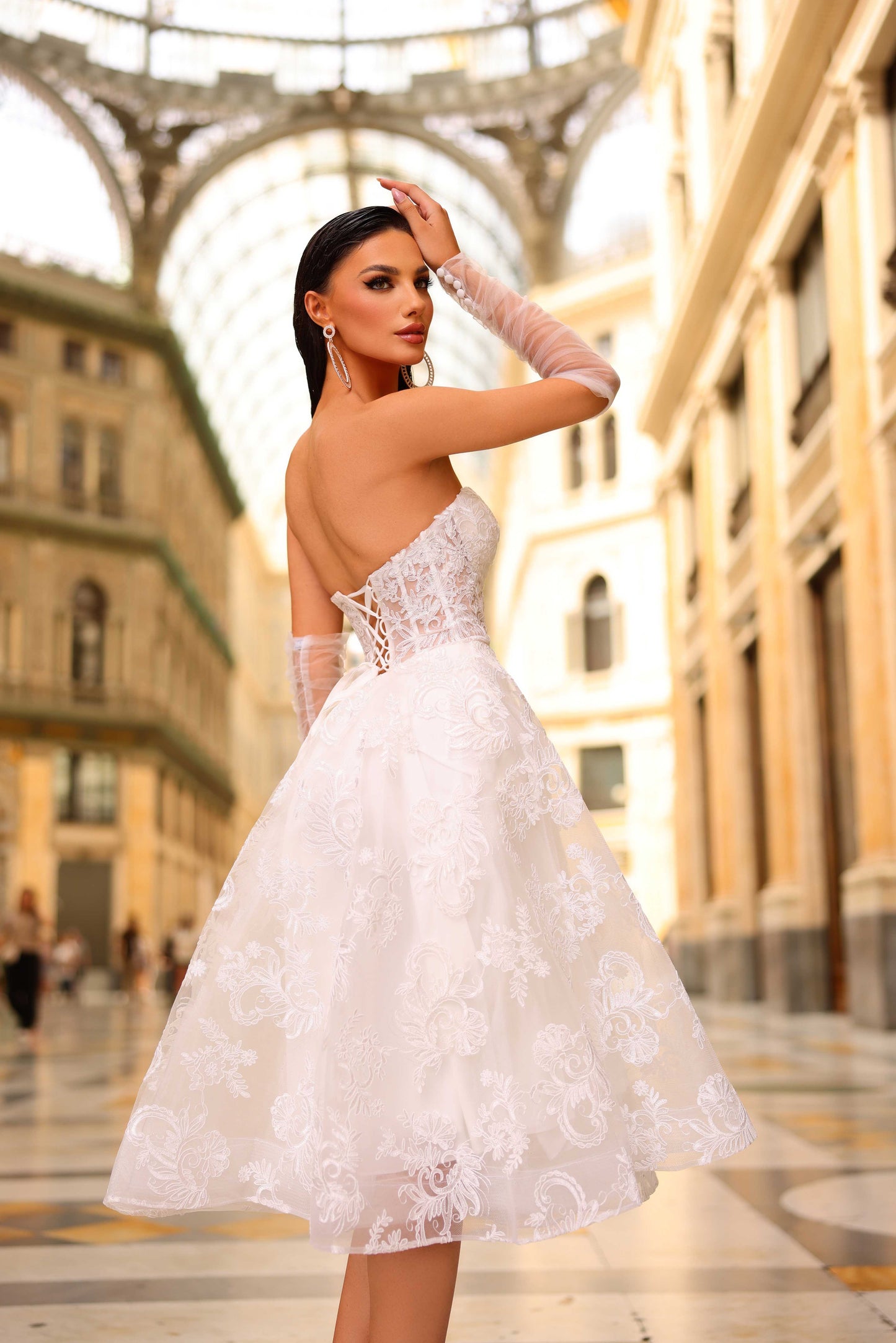 Woman in a white lace dress standing in an urban setting with architectural elements.