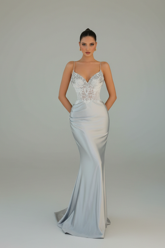 A woman modeling a floor-length satin evening gown with a lace-embellished sweetheart bodice and thin spaghetti straps, photographed against a seamless light gray studio background.