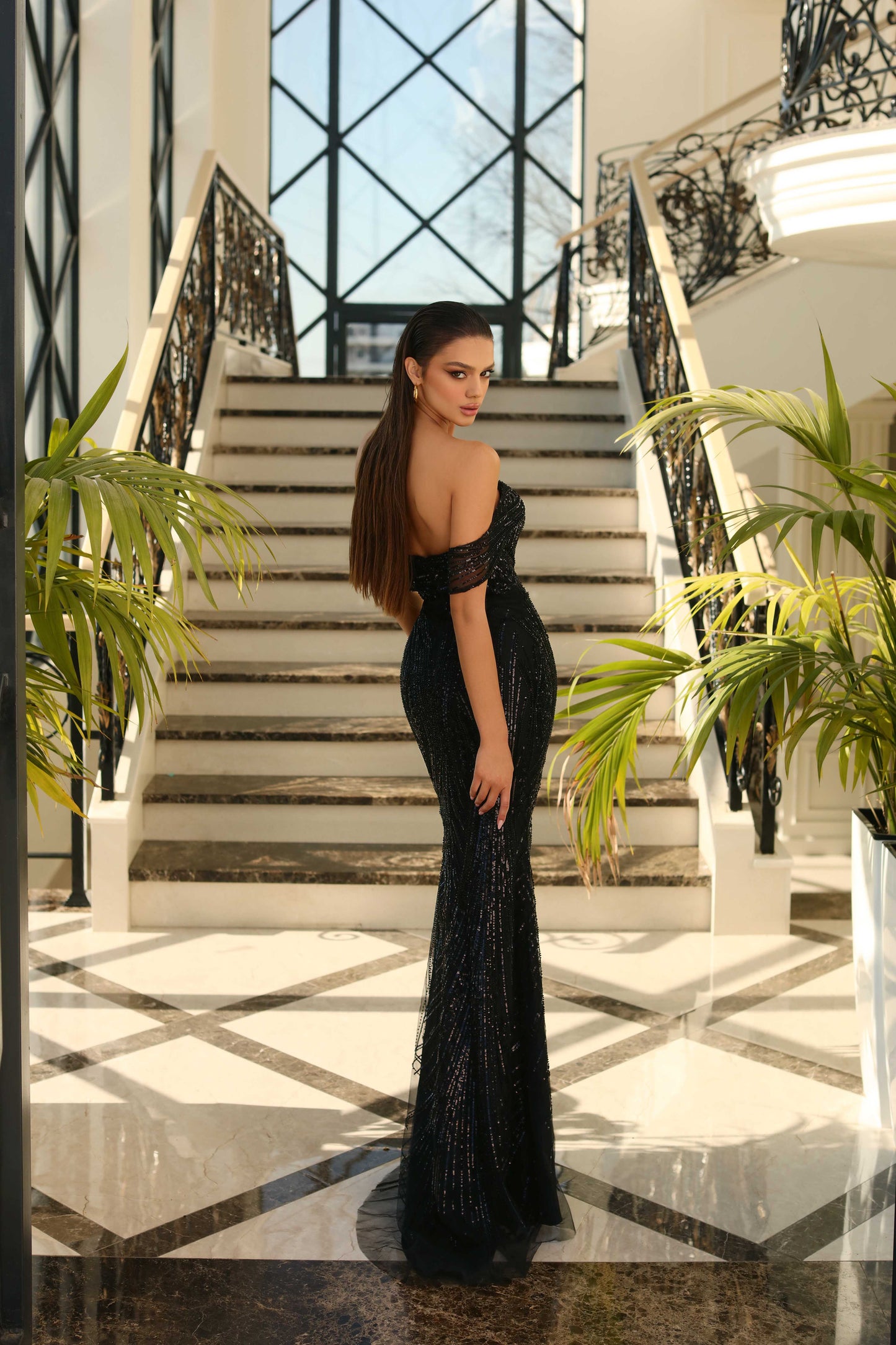 Woman in a black evening gown standing in a grand staircase with decorative railings and plants.