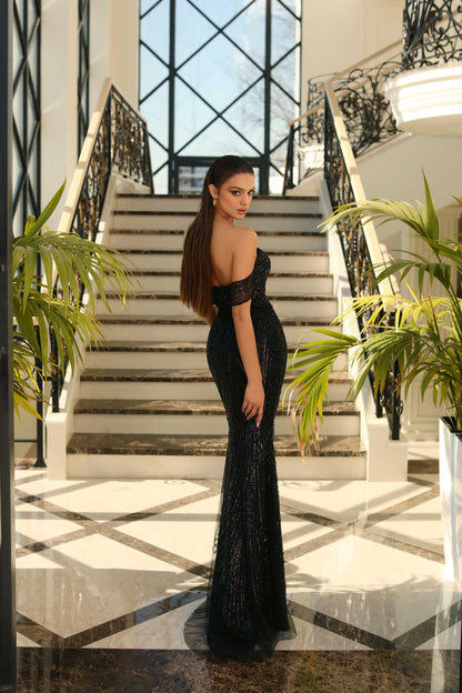 Woman in a black evening gown standing in a grand staircase with decorative railings and plants.
