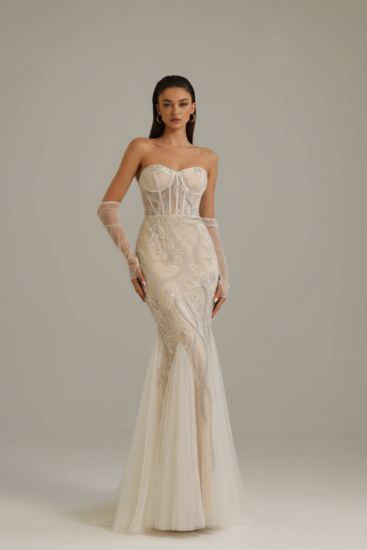 A woman modeling a strapless ivory wedding gown with long sheer sleeves, a fitted lace bodice, and a flowing tulle mermaid skirt against a plain light gray studio background.