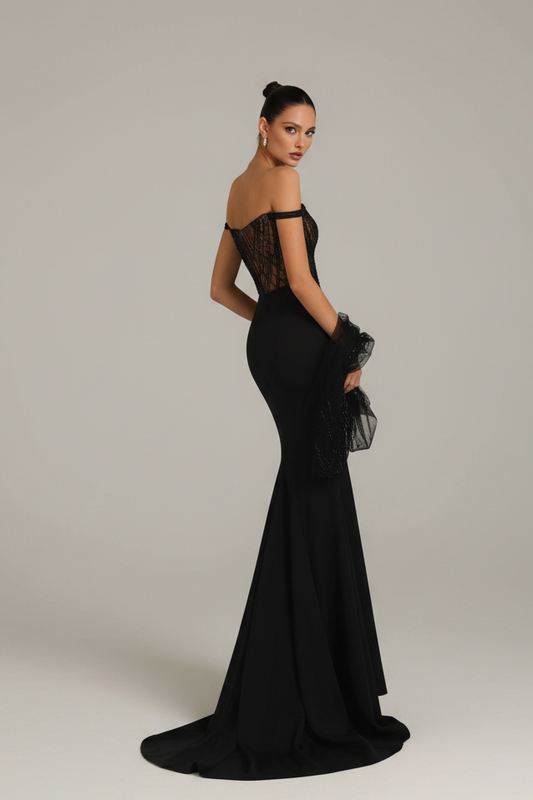A woman modeling a floor-length black off-the-shoulder evening gown with sheer lace detailing on the bodice, photographed against a plain light gray studio background.