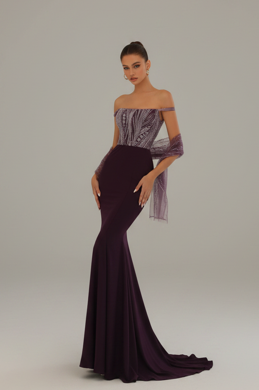 A model wearing a floor-length off-the-shoulder evening gown with a deep V-neckline, a structured bodice, sheer draped sleeves, and a long train, photographed against a neutral light gray studio background.