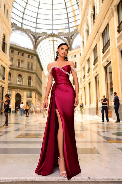 Woman in a burgundy evening gown with a high slit standing in an elegant indoor setting.