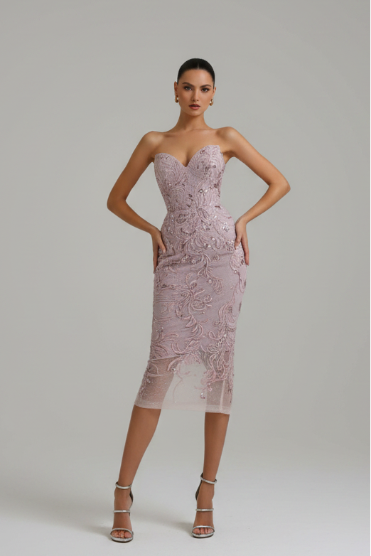 A model wearing a strapless, fitted midi dress in a dusty pink lace and mesh pattern, standing against a neutral light gray studio background.