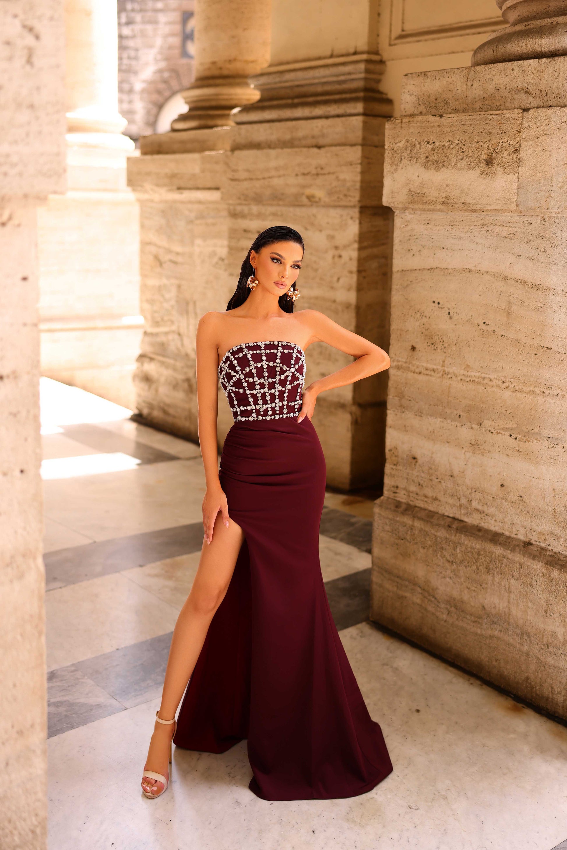 Woman in a burgundy evening gown with a high slit standing in an architectural setting.