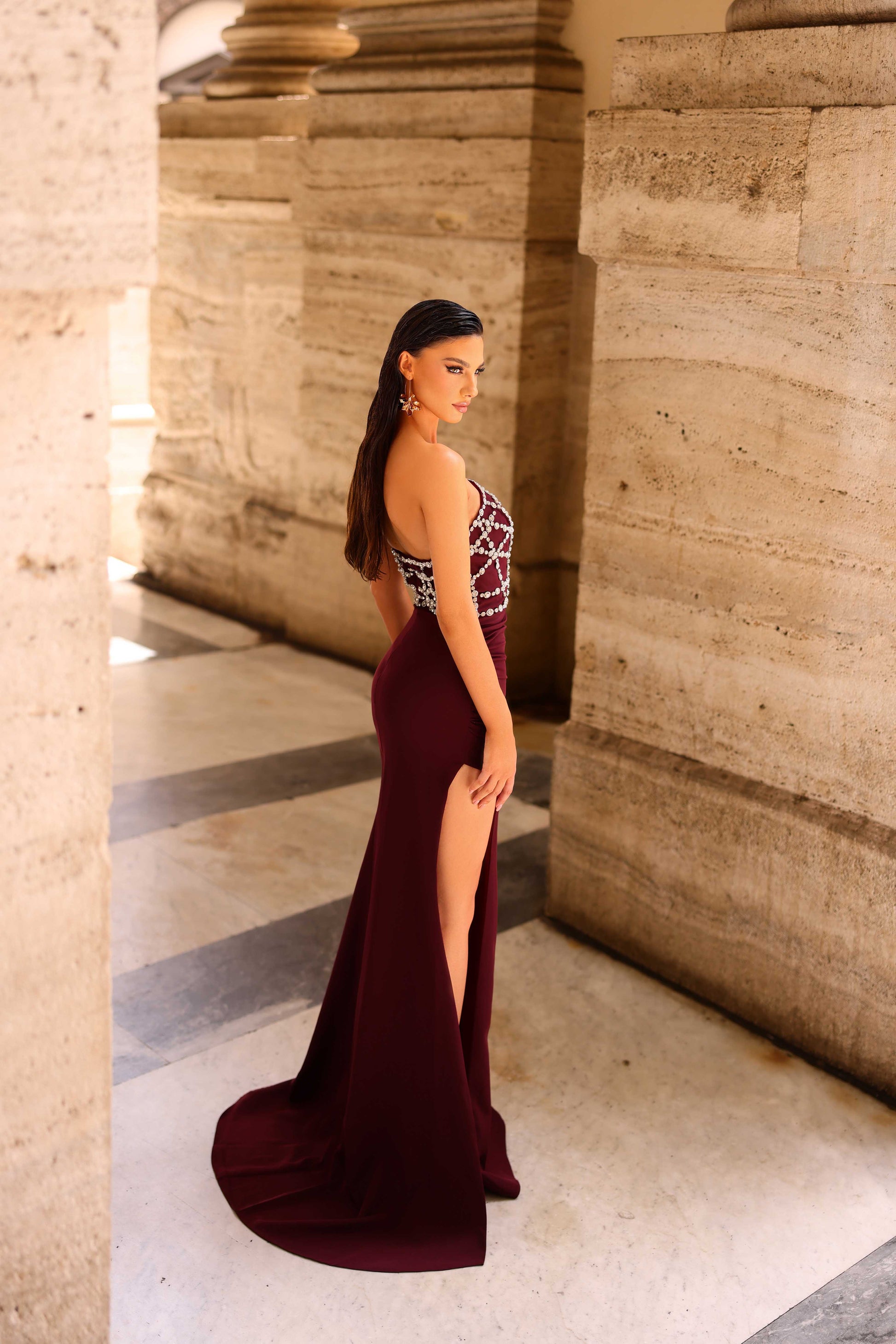 Woman in a burgundy evening gown with a high slit standing against stone columns.