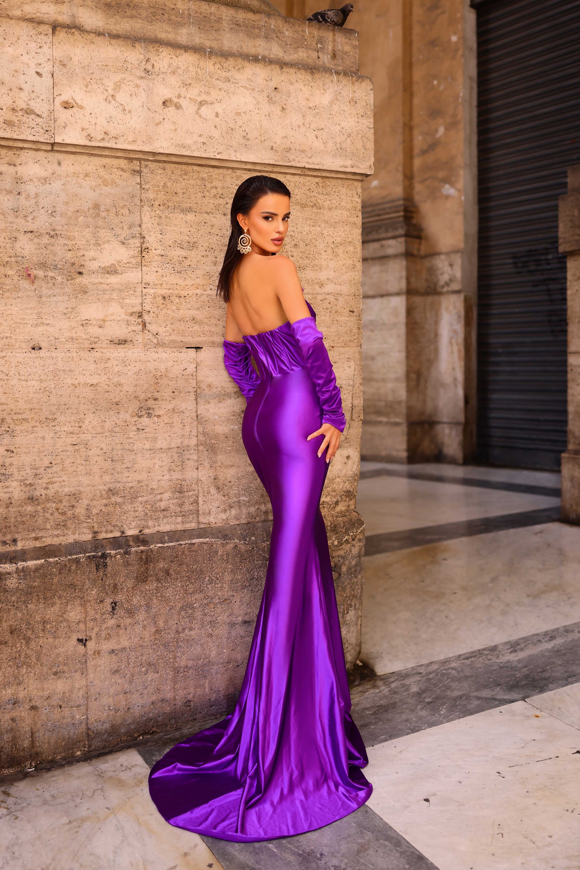 Woman in a purple evening gown standing against a stone wall.