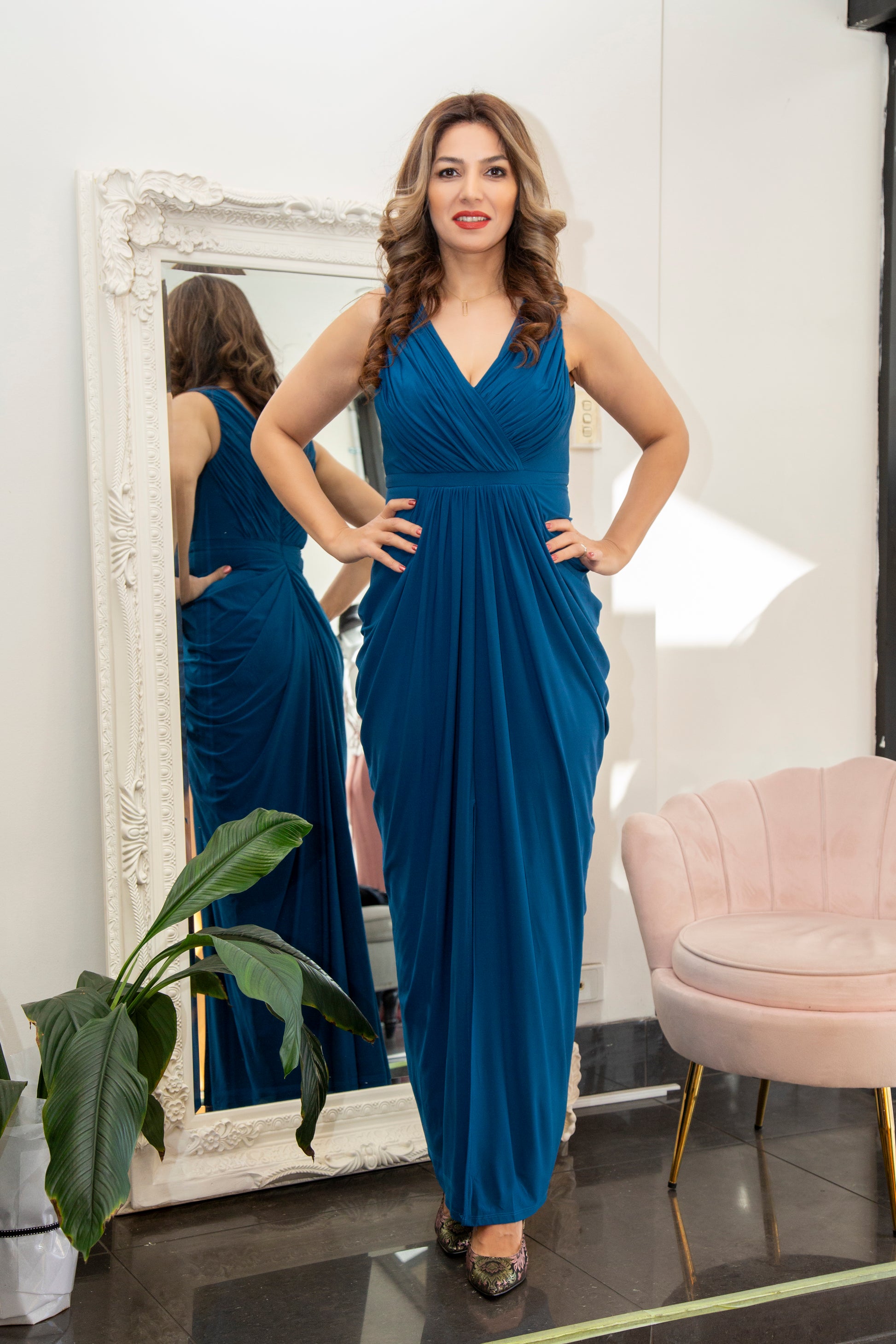 Woman in a blue dress standing in front of a mirror in a room with a pink chair and plant.