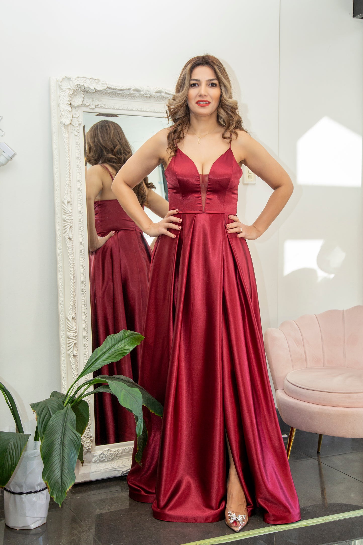 Woman in a red dress standing in front of a mirror in a room with a plant and pink chair.