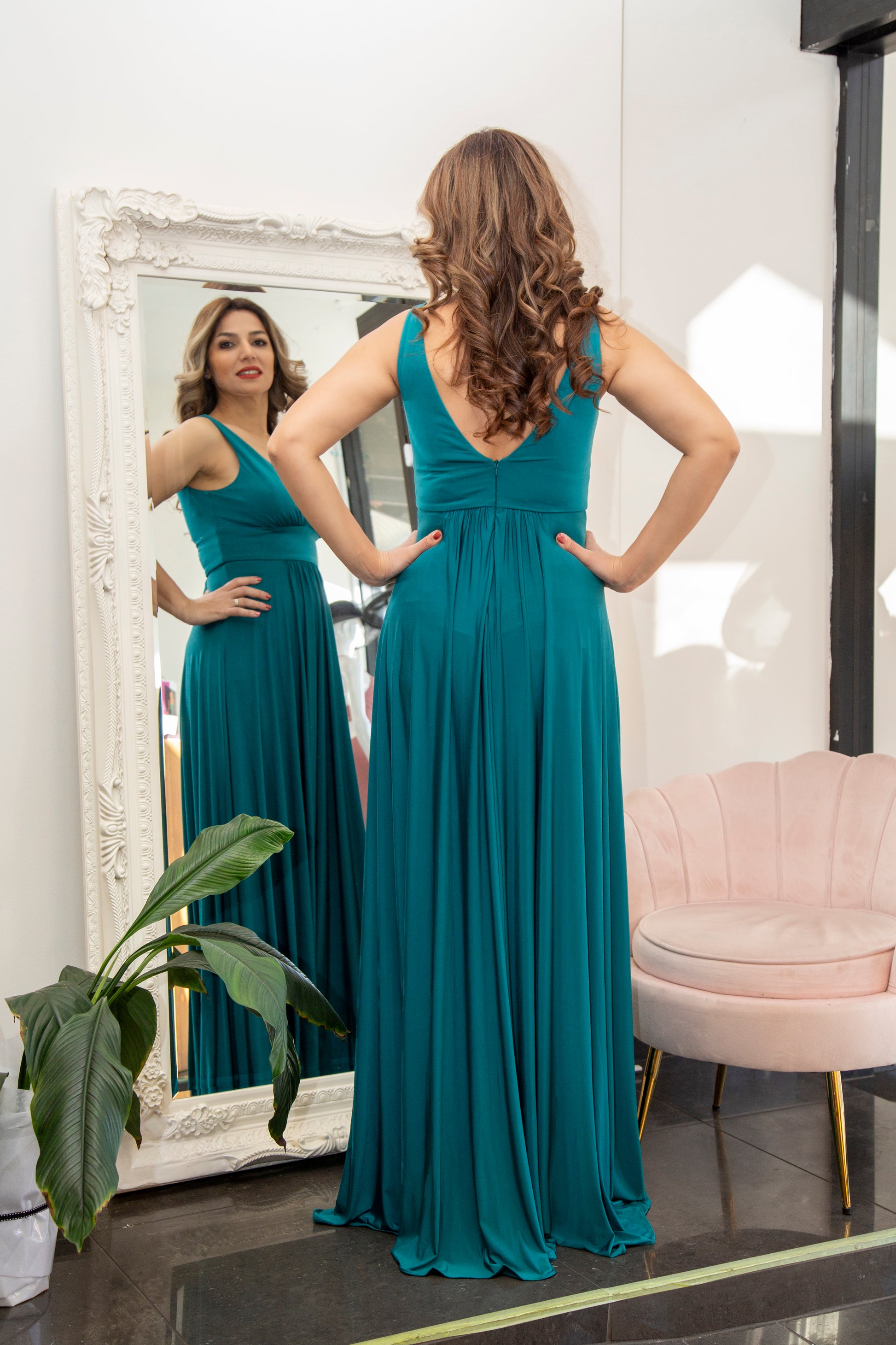 Woman in a teal dress standing in front of a mirror in a room with a pink chair and plant.