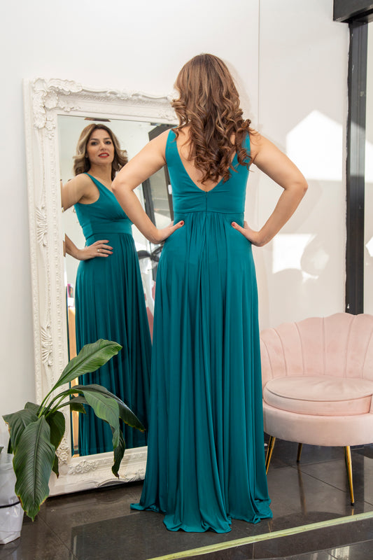Woman in a teal dress standing in front of a mirror in a room with a pink chair and plant.