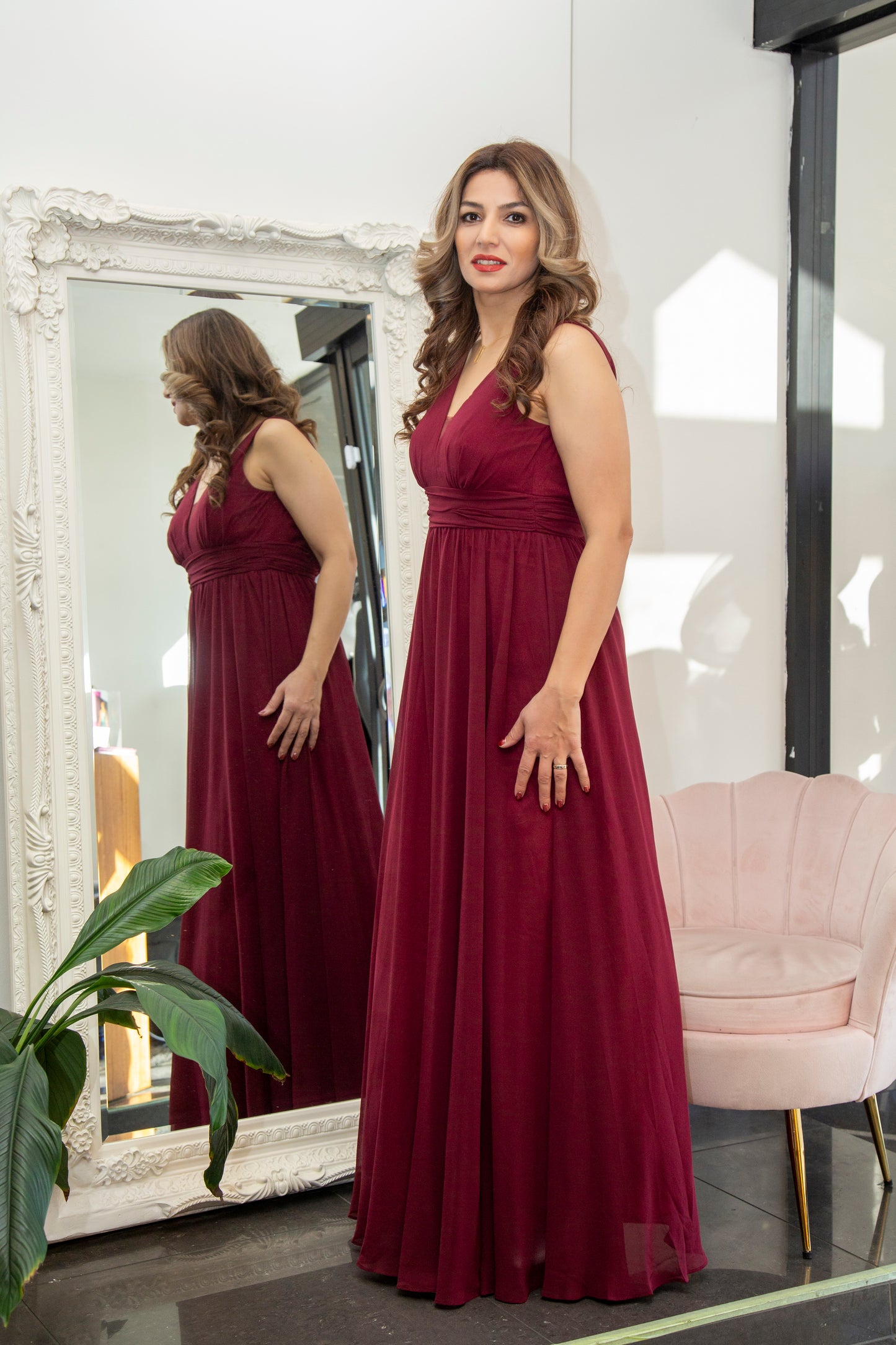 Woman in a burgundy dress standing in front of a mirror in a room with a chair and plant.