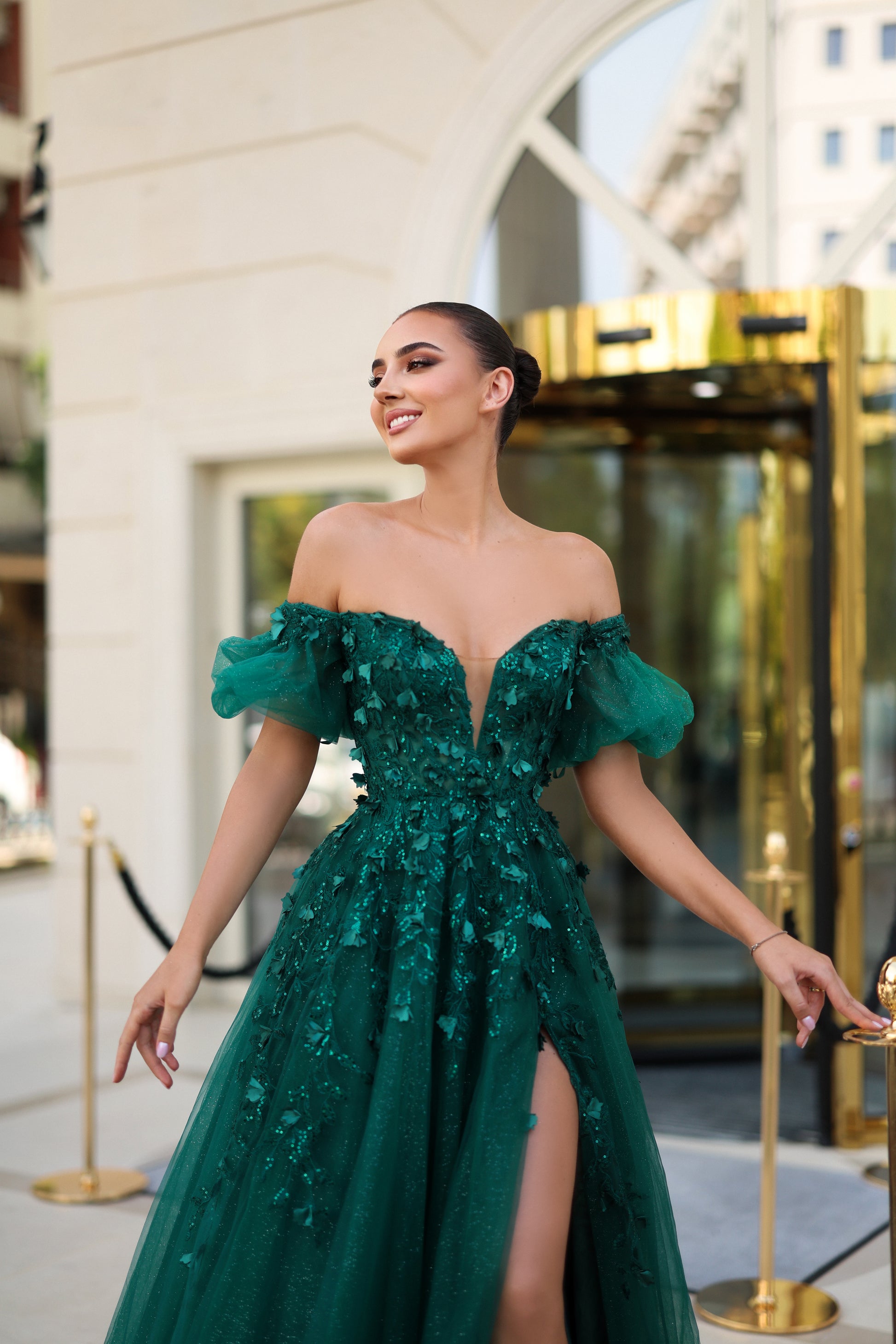 Woman in a green off-shoulder gown with floral details standing in front of a building entrance.