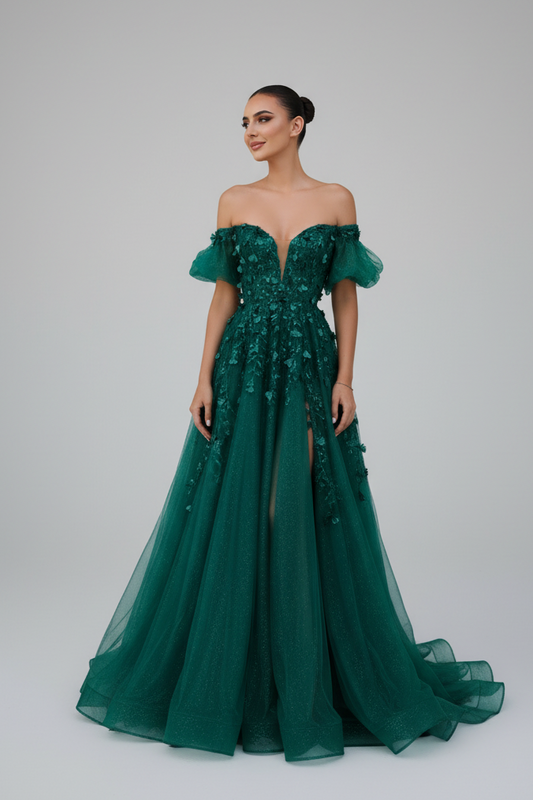A woman modeling an off-the-shoulder emerald green evening gown with intricate lace applique on the bodice and a flowing tulle skirt, set against a plain light gray studio background.