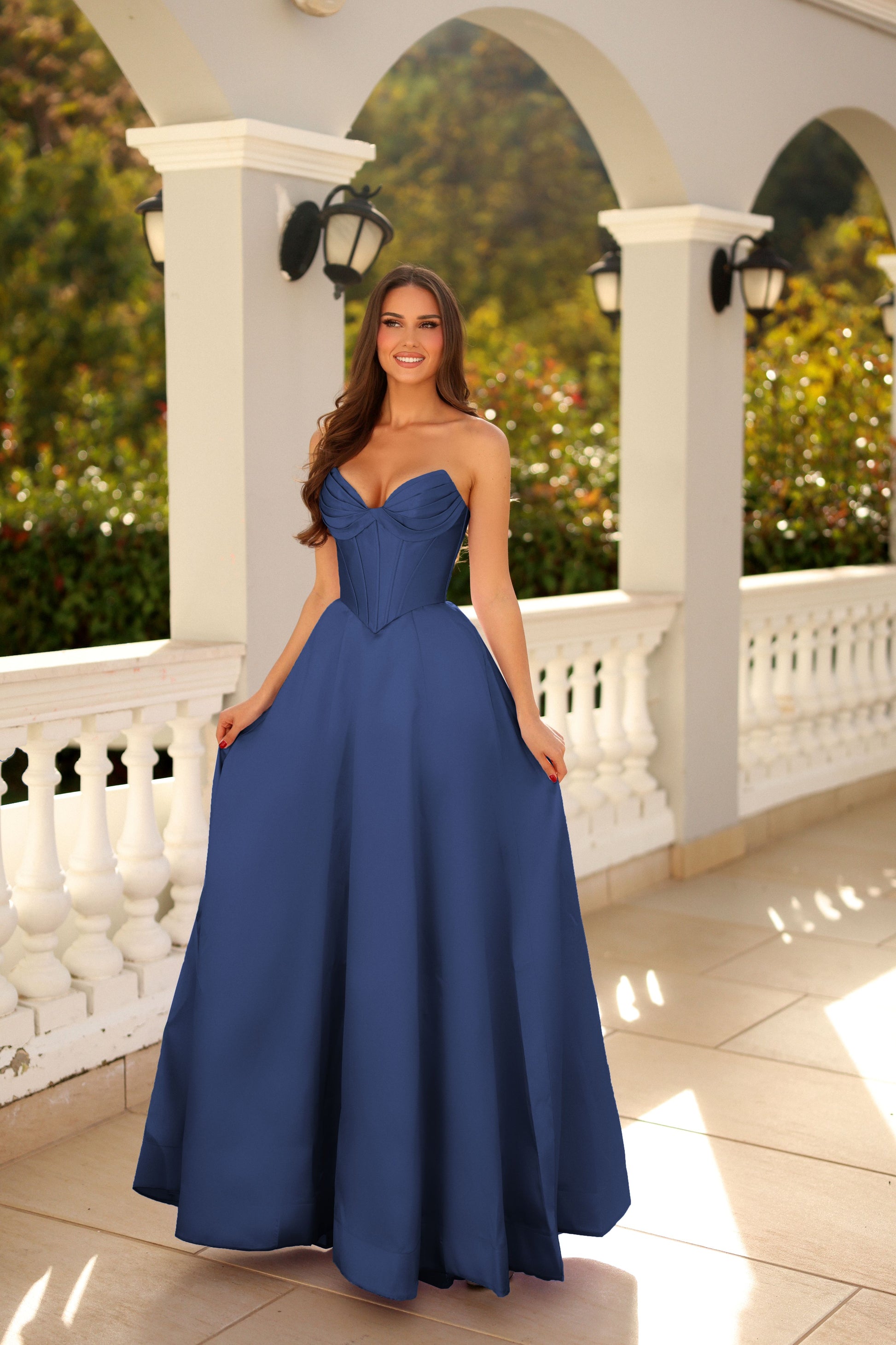 Woman in a blue strapless gown standing on a balcony with white columns and greenery.
