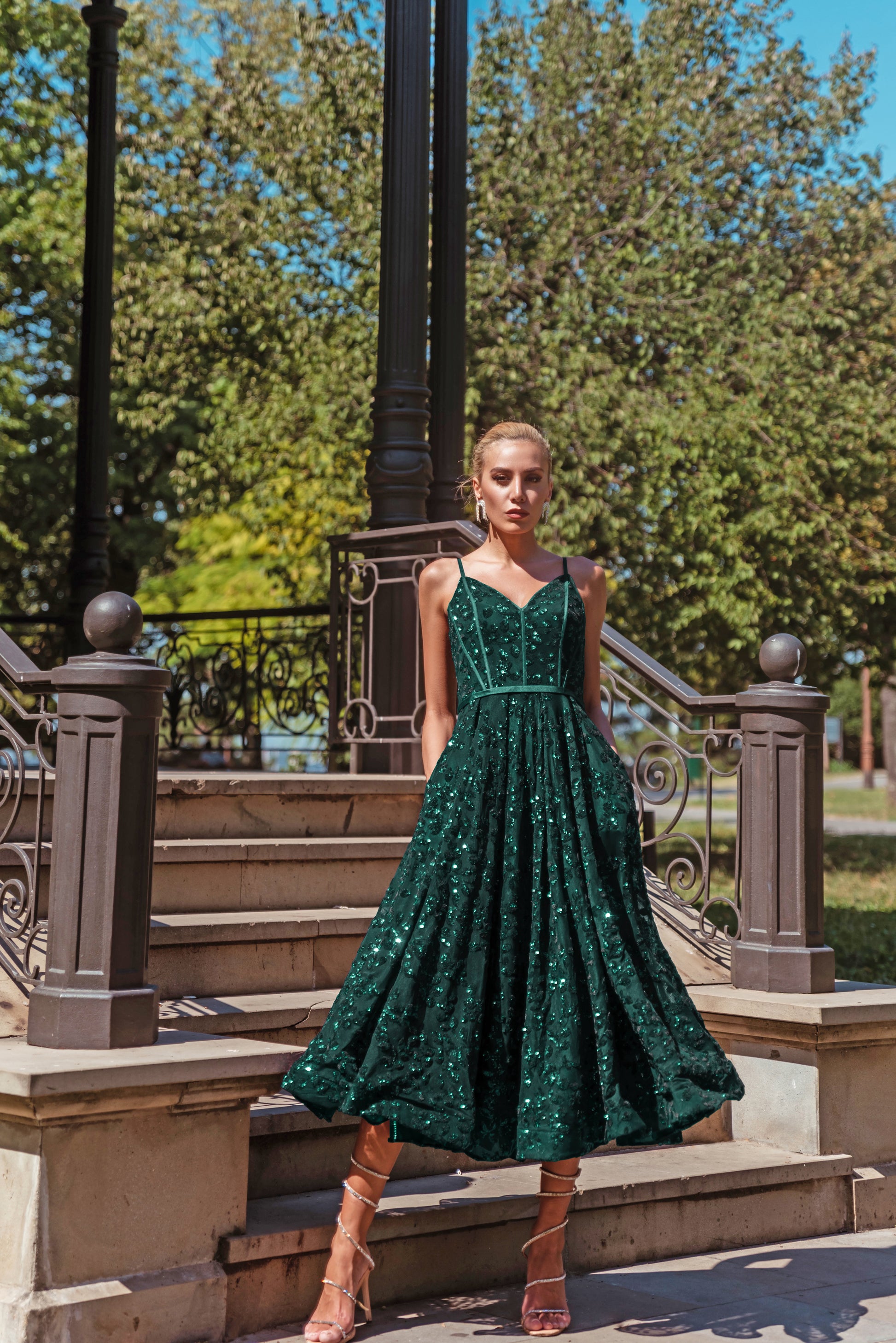 Woman in a green sequined dress standing on steps outdoors with trees in the background