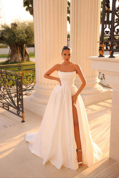 Woman in a white strapless gown with a high slit standing in an elegant outdoor setting.